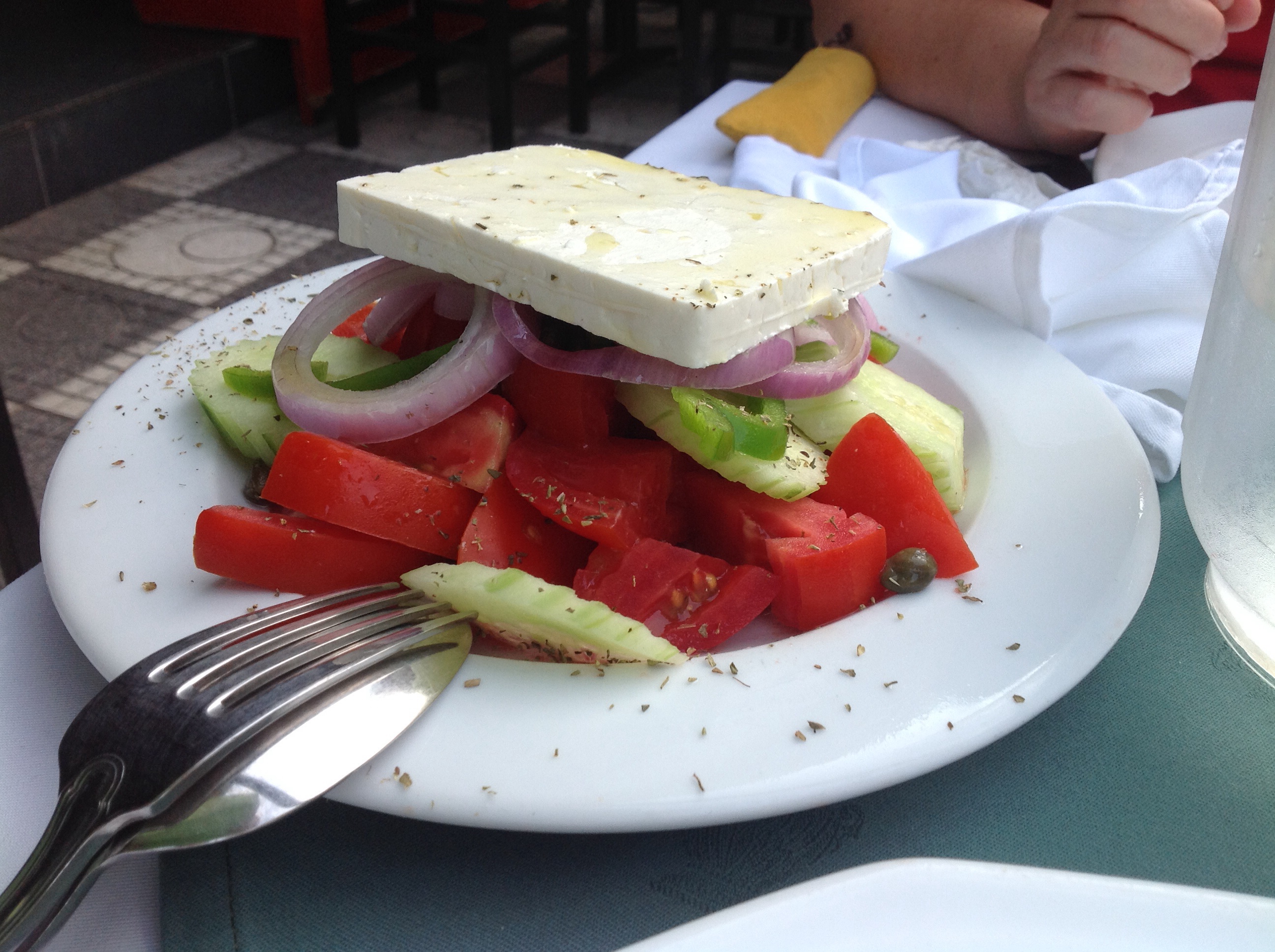 A Greek village salad of tomato, onion cucumber and a large slab of feta cheese on white plate with fork and spoon overturned
